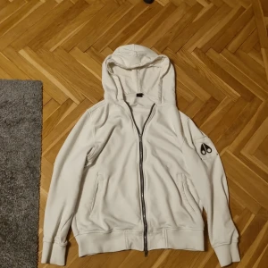 Moose Knuckles Snyider Full-Zip - Säljer en stilren vit hoodie från Moose Knuckles med dragkedja och huva. Tröjan har ett unikt märke på ärmen och ribbade muddar vid ärmslut och nederkant. Perfekt för en avslappnad stil.