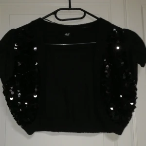 Svart bolerotopp från H&M - Snygg svart bolerotopp från H&M med glittriga paljetter på framsidan. Perfekt för att lägga till lite glamour till din outfit. Kortärmad design som passar bra till festliga tillfällen.