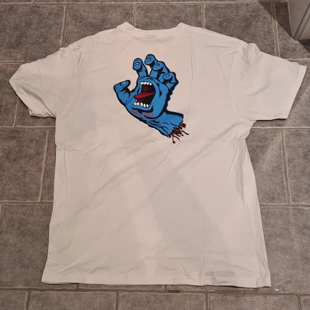 Säljer en vit t-shirt med ett coolt blått tryck av en skrikande hand på både fram- och baksidan. Perfekt för en avslappnad stil. Märke Santa Cruz. Knappt använd. Storlek L. T-paidat.
