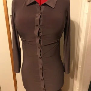 Brun klänning från Tille - Snygg brun klänning från Tille med långärmad design och knappar framtill. Perfekt för en stilren look. Tillverkad i Turkiet.