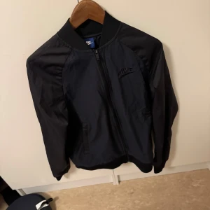 Svart bomberjacka från Nike - Snygg svart bomberjacka från Nike med dragkedja framtill. Jackan har en klassisk design med ribbad krage och muddar. Perfekt för en stilren look.