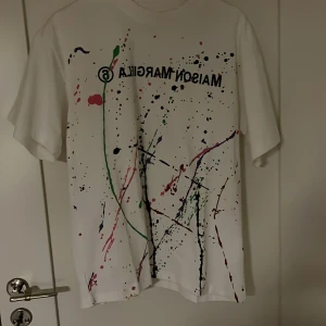 Vit t-shirt från Maison Margiela - Snygg vit t-shirt från Maison Margiela med färgstänk i grönt, rött och svart. Den har en avslappnad passform och korta ärmar. Perfekt för en trendig och avslappnad look.Den har en liten fläck på bak vänster axel. Den är från 🐼