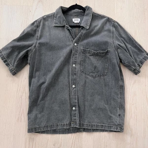 Grå jeansskjorta från Weekday - Säljer en grå jeansskjorta från Weekday i storlek M. Skjortan har korta ärmar och knappar framtill samt en bröstficka.