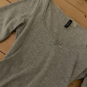 Grå topp från H&M Divided - Jättesöt grå tröja från H&M. Den har en liten rosett, spets i kanten och fint mönster (se sisha bilden). I bra skick, nästan aldrig använd. 