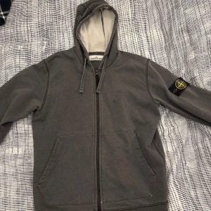 Grå hoodie från Stone Island - Säljer en stilren grå hoodie från Stone Island med dragkedja och huva. Tröjan har en ikonisk logga på ärmen och praktiska fickor framtill.  storlek M men passar S