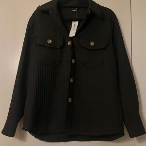 Svart skjort/jacka från Gina Tricot - Snygg svart skjorta från Gina Tricot med knappar framtill och två bröstfickor. Skjortan har en klassisk krage och långa ärmar, perfekt för en stilren look.
