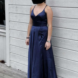 Mörkblå långklänning med snörning - Elegant mörkblå långklänning i satin med v-ringad hals och smala axelband. Klänningen har en stilren slits framtill och en vacker snörning i ryggen som ger en perfekt passform. Perfekt för festliga tillfällen.
