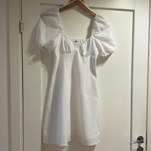 Vit klänning  - Vit kort klänning med struktur i tyget. Jättefin med detalj fram vid brösten, liten ballongärm och smock i ryggen för fin passform. Endast använd 1 gång men lappen bortklippt. Troligen strl xs. 