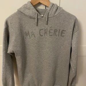 Grå hoodie från Gina Tricot - Säljer en stilren grå hoodie från Gina Tricot med texten 'Ma Chérie' i glittriga stenar på framsidan. Tröjan har en klassisk huva med dragsko och långa ärmar. Perfekt för en avslappnad look.