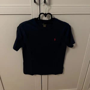 Mörkblå t-shirt med röd logga - Snygg mörkblå t-shirt från Ralph Lauren med en liten röd broderad logga på bröstet. Perfekt för en stilren och avslappnad look.