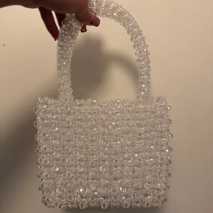 Pärlhandväska - Snygg och unik handväska helt täckt av transparenta pärlor. Perfekt för att ge en glittrig touch till din outfit. Väskan har ett handtag och är i en kompakt storlek, vilket gör den lätt att bära med sig.