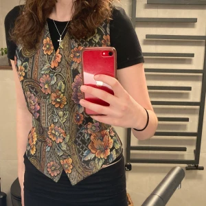 Blommig väst med knappar - Säljer en unik väst med ett färgglatt blommönster i orange, rosa och grönt. Västen har knappar framtill och en elegant passform. Perfekt för att ge din outfit en vintagekänsla.