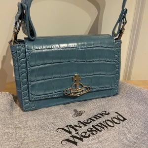 Blå axelväska från Vivienne Westwood - Snygg blå axelväska i krokodilpräglat skinn från Vivienne Westwood. Väskan har ett ikoniskt metallspänne framtill och en justerbar axelrem. Perfekt för att ge din outfit en lyxig touch.