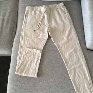 Beige byxor från Knowledge Cotton Apparel - Snygga beige byxor från Knowledge Cotton Apparel i en skön blandning av bomull och linne. Byxorna har en avslappnad passform med snörning i midjan och praktiska fickor. Perfekta för en stilren och bekväm look. Linnebyxor.