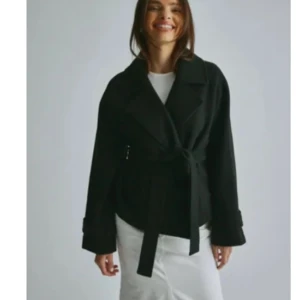 Svart kappa med knytband - Elegant svart kappa med knytband i midjan. Perfekt för en stilren look. Långärmad design med klassisk krage. Har använts fåtals gånger och kommer ej till användning nypriss 599