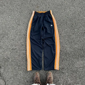 Svarta träningsbyxor från Adidas - Snygga svarta träningsbyxor från Adidas med orange och vita ränder längs sidorna. Byxorna har en elastisk midja för extra komfort och är perfekta för sportiga aktiviteter. Adidas-loggan är diskret placerad på framsidan.