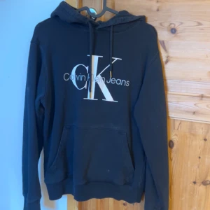 Svart hoodie från Calvin Klein - Säljer en svart hoodie från Calvin Klein med det klassiska CK-logotypen på bröstet. Tröjan har en justerbar huva med dragsko och en stor magficka framtill. Perfekt för en avslappnad stil.
