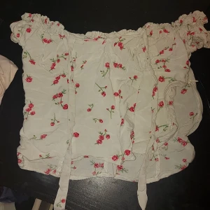 Blommig offshoulder topp från Bershka - Söt offshoulder topp från Bershka i vitt med röda blommor. Toppen har en rynkad design och knytband för extra detaljer. Perfekt för en somrig look!