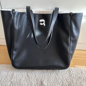 Karl lagerfeldt väska - Snygg svart shoppingväska med en cool ikon på framsidan. Väskan har en vit, fluffig insida och svarta handtag. Perfekt för att bära med sig allt man behöver under dagen.