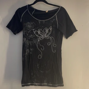 Svart kortärmad topp med strassdetaljer - Snygg svart kortärmad topp med glittrande strassdetaljer i form av en fjäril på framsidan. Toppen har vita sömmar som ger en fin kontrast och en rund halsringning. Perfekt för att lägga till lite bling i vardagen! nån ynka sten har trillat av💗annars nyskick
