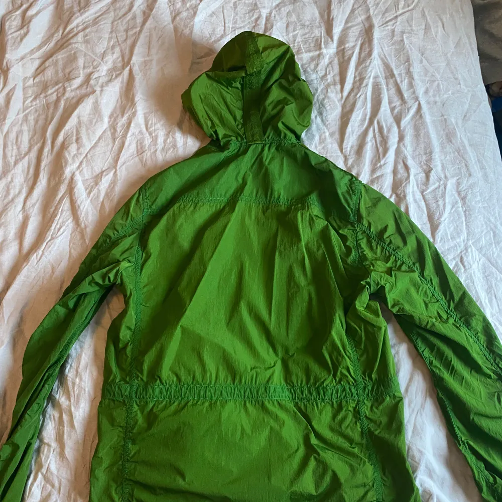Säljer en grön vindjacka från C.P. Company. Jackan har en huva och dragkedja framtill. Den är utrustad med flera fickor med knappar och dragkedjor för praktisk förvaring. Perfekt för blåsiga dagar.. Takit.