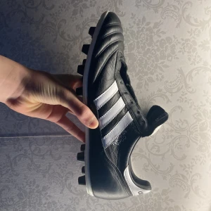 Svarta fotbollsskor från Adidas, COPA MUNDIAL. - Klassiska svarta fotbollsskor från Adidas med vita detaljer och de ikoniska tre ränderna på sidan. Skorna har snörning och dobbar för bra grepp på planen. Perfekta för fotbollsspelare som vill ha stil och funktion.