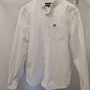 Vit skjorta från Lyle & Scott - Säljer en stilren vit skjorta från Lyle & Scott med klassisk krage och knappstängning. Skjortan har en bröstficka med det ikoniska broderade logotypen i gult och svart. Perfekt för en snygg och avslappnad look.