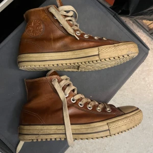 Bruna skinnsneakers från Converse - Snygga bruna skinnsneakers från Converse med klassisk hög modell och snörning. Skorna har en robust sula och det ikoniska Converse-märket på sidan. Perfekta för en stilren och avslappnad look.