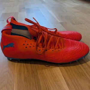 Röda fotbollsskor från Puma - Snygga röda fotbollsskor från Puma med blå detaljer och snörning. Skorna har en mönstrad yta och dobbar för bra grepp på planen. Perfekta för fotbollsspelare som vill sticka ut.