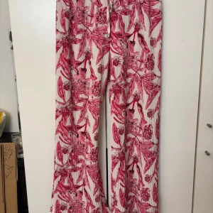 Blommiga byxor med vida ben - Snygga byxor med ett iögonfallande blommigt mönster i rosa och rött på en vit bakgrund. Byxorna har vida ben och en bekväm passform, perfekta för en somrig look.