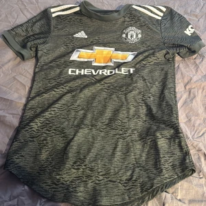 Grön Manchester United fotbollströja - Säljer en grön Manchester United fotbollströja från Adidas med korta ärmar. Tröjan har ett subtilt mönster och klubbens emblem på bröstet. Den har även sponsorloggan Chevrolet på framsidan och spelarnamnet 'Ronaldo' med nummer 7 på baksidan.