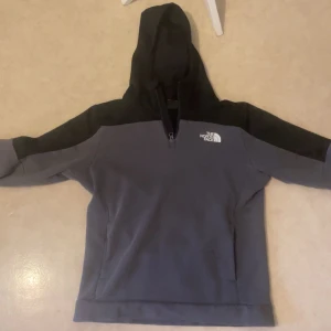 Grå och svart hoodie från The North Face - Säljer en snygg grå och svart hoodie från The North Face. Den har en klassisk design med huva och dragkedja vid halsen. Perfekt för kyliga dagar och har en bekväm passform.