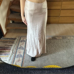 Beige långkjol - Säljer en elegant beige långkjol med en slät och glansig yta. Kjolen har en figurnära passform och är perfekt för en stilren look. Den är tillverkad i ett lätt material som ger ett fint fall.