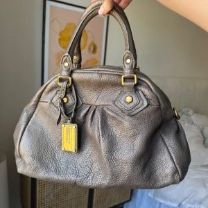 Väska från Marc Jacobs - Snygg gråbeige väska från Marc Jacobs. Väskan har guldfärgade detaljer och en unik innerfoder med textmönster. Passar perfekt till skolan och klär upp en vardagsoutfit! Knappt använd. Älskar den!