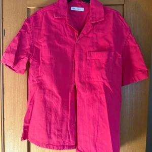 Rosa kortärmad skjorta från Zara - Snygg rosa skjorta från Zara med korta ärmar och knappar framtill. Perfekt för en färgglad look. Skjortan har en bröstficka som ger en extra touch. Passar bra till både jeans och shorts.