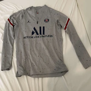 Grå långärmad fotbollströja från Jordan x PSG - Säljer en grå långärmad fotbollströja från Jordan i samarbete med PSG. Tröjan har ett diskret stänkmönster och detaljer i rött och blått på ärmarna. Den har en halv dragkedja framtill och PSG-loggan på bröstet. Den är båda för tjejer och killar och är äkta knappt använd.  Priset kan diskuteras 