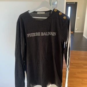 Mörkgrå långärmad tröja från Pierre Balmain - Säljer en stilren mörkgrå långärmad tröja från Pierre Balmain. Tröjan har dekorativa knappar på axeln och ett tryck med märkesnamnet på framsidan. Tillverkad i en mjuk bomullsblandning för extra komfort. Perfekt för en avslappnad men ändå elegant look.