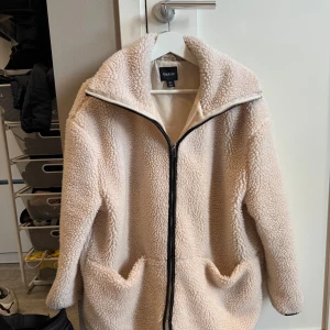 Beige teddyjacka från Boohoo - Mysig beige teddyjacka från Boohoo med dragkedja framtill. Jackan har en fluffig textur och långa ärmar, perfekt för kyliga dagar. Den har en avslappnad passform och är tillverkad i syntetmaterial.