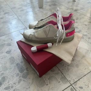 Vita sneakers från Valentino - Snygga vita sneakers från Valentino med rosa detaljer och perforerat mönster på sidorna. Skorna har snörning och en klassisk design med en modern twist. Perfekta för att ge en färgklick till din outfit.