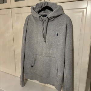 Grå hoodie från Polo Ralph Lauren - Säljer en stilren grå hoodie från Polo Ralph Lauren med den klassiska loggan broderad i mörkblått på bröstet. Tröjan har en bekväm passform med känguruficka och justerbar huva med snören. Mycket bra skick, storlek M men passar även S!