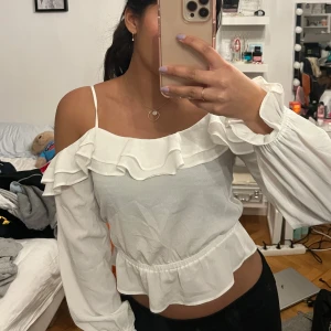 Vit blus med volanger - Säljer en söt vit blus med volanger och smala axelband. Blusen har långa ärmar med elastiska ärmslut och en luftig passform. Perfekt för en somrig look!
