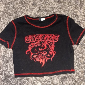 Svart croppad t-shirt med röd text - Säljer en svart croppad t-shirt med röd text och ormmotiv på framsidan. Köpt på HM för några år sedan. Använd ett fåtal gånger. Storlek S men ganska liten i storleken.
