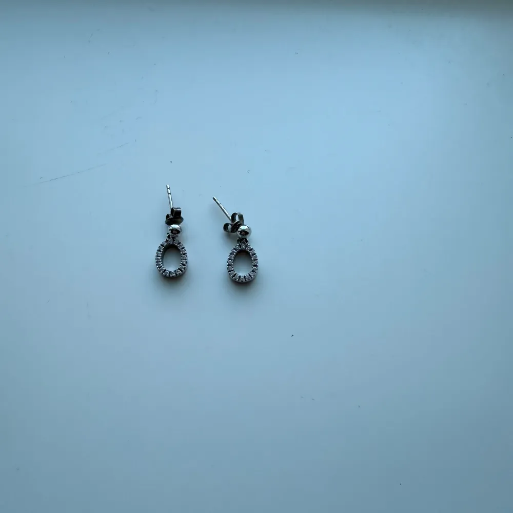 Eleganta silverfärgade örhängen med glittriga stenar i en oval form. Perfekta för att ge en touch av glamour till din outfit. De har en enkel stiftstängning som gör dem lätta att bära.. Asusteet.