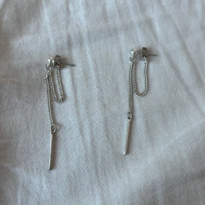 Silvriga örhängen med kedja - Eleganta silvriga örhängen med en tunn kedja och stavdetalj. Perfekta för att ge en stilren touch till din outfit. Lätta och bekväma att bära hela dagen.