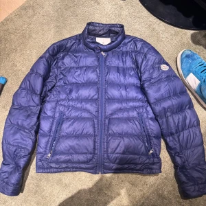 Blå dunjacka från Moncler - Säljer en snygg blå jacka från Moncler. Jackan har en quiltad design med dragkedja framtill och två praktiska fickor. Perfekt för höst/vår och ger en stilren look.