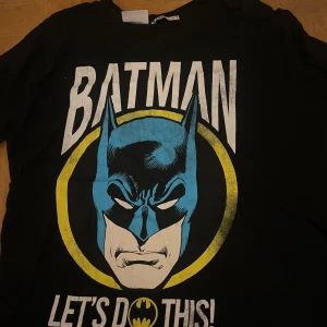 Svart Batman t-shirt från FB Sister - Cool svart t-shirt med Batman-tryck från FB Sister. T-shirten har ett stort tryck av Batman i blått och gult med texten 'Let's Do This!'. Perfekt för alla Batman-fans! Köpt från new yorker