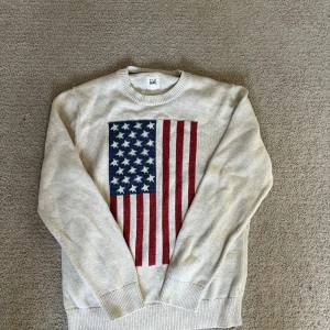 Beige tröja med amerikansk flagga - Säljer en beige tröja från GAP med en stor amerikansk flagga på framsidan. Tröjan har långa ärmar och ribbade muddar vid ärmslut och nederkant. Perfekt för en avslappnad stil!