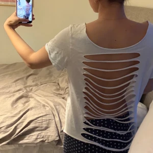 Vit t-shirt med öppen rygg - Säljer en vit t-shirt med en unik design på baksidan där tyget är skuret i ett mönster som skapar en öppen rygg. T-shirten har en avslappnad passform och korta ärmar, perfekt för en casual look.
