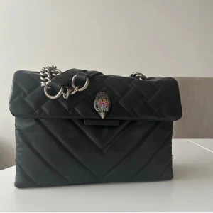 Svart Kurt Geiger quiltad axelväska med kedja - Snygg svart axelväska med quiltat mönster och en elegant kedja som axelrem. Väskan har en unik detalj på framsidan och en rund logotyp på baksidan. Perfekt för att ge en stilren touch till din outfit.