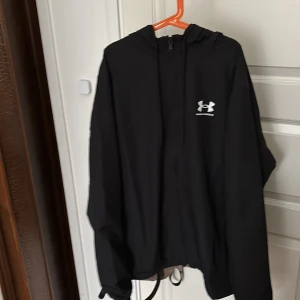 Svart vindjacka från Under Armour - Säljer en svart vindjacka från Under Armour med huva och dragkedja. Jackan har en enkel design med logotyp på bröstet och är perfekt för blåsiga dagar. Den är tillverkad i ett lätt syntetmaterial och har långa ärmar.